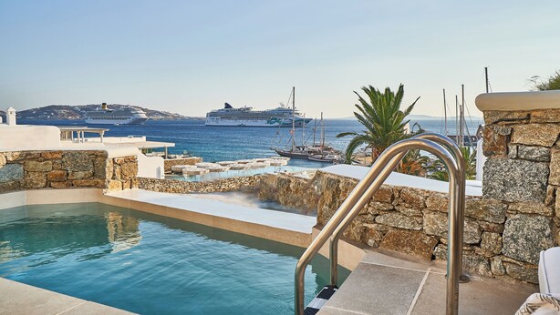 A swimming pool with stone walls and metal railings, overlooking a marina with boats and a cruise ship. - Aqua Mykonos Suites - Adults Only (Mykonos)