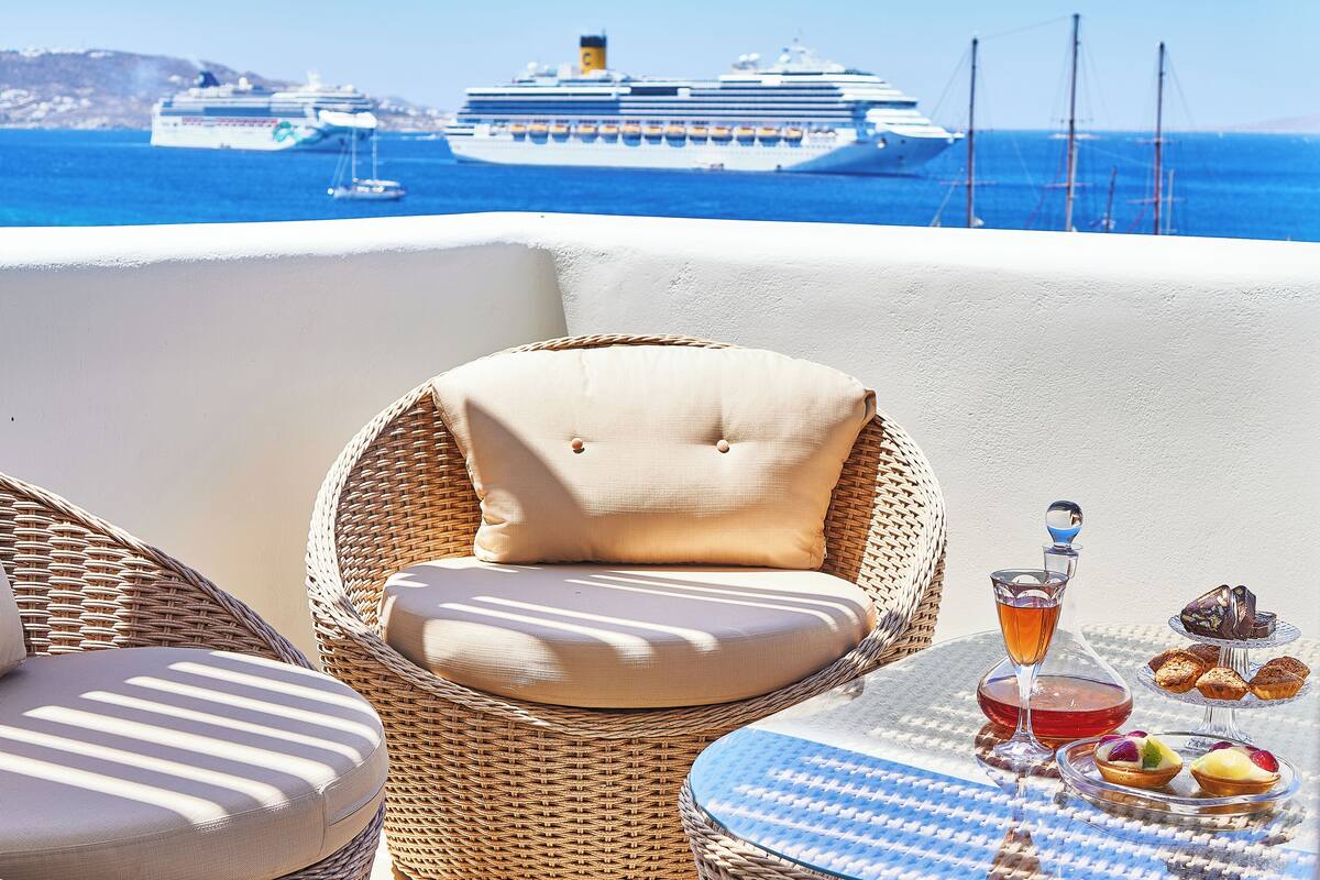 A wicker chair with a cushion, a table with a glass of drink and pastries, and a cruise ship in the background.