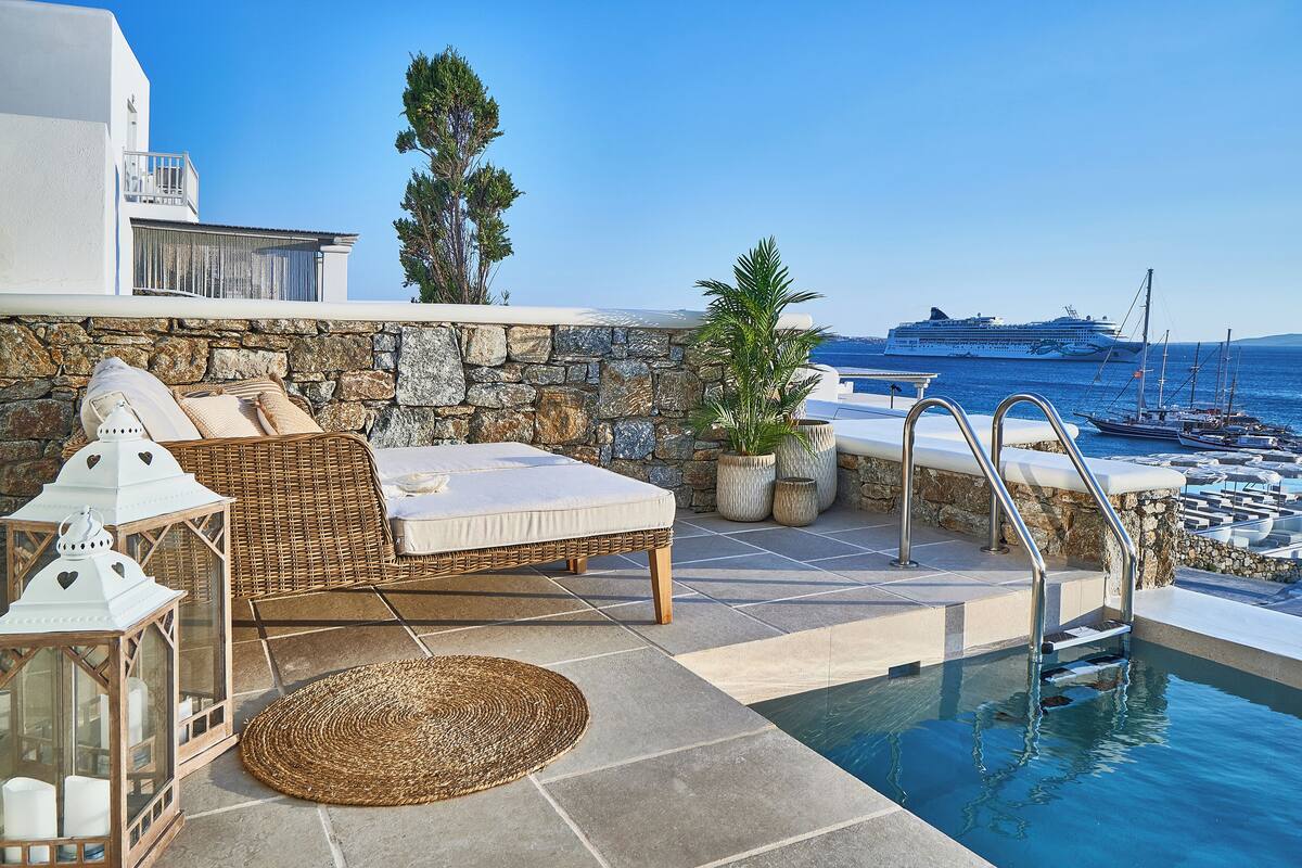 A poolside area with a lounge chair, a stone wall, and a view of the sea and a cruise ship.
