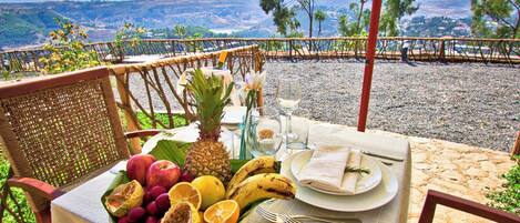 A table set with fruits, glasses, and cutlery on a terrace overlooking a scenic landscape.
