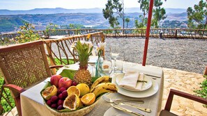 Mesa posta com frutas, copos e talheres em um terraço com vista para uma paisagem deslumbrante.