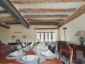 A dining room with a long table set for a meal, wooden beams on the ceiling, and a window providing natural light.