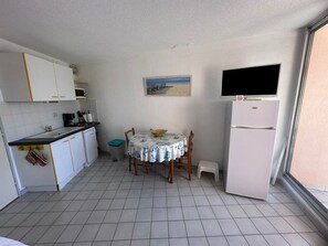 Une cuisine compacte avec des meubles blancs, un sol carrelé, une petite table à manger avec des chaises, un réfrigérateur et un téléviseur fixé au mur.