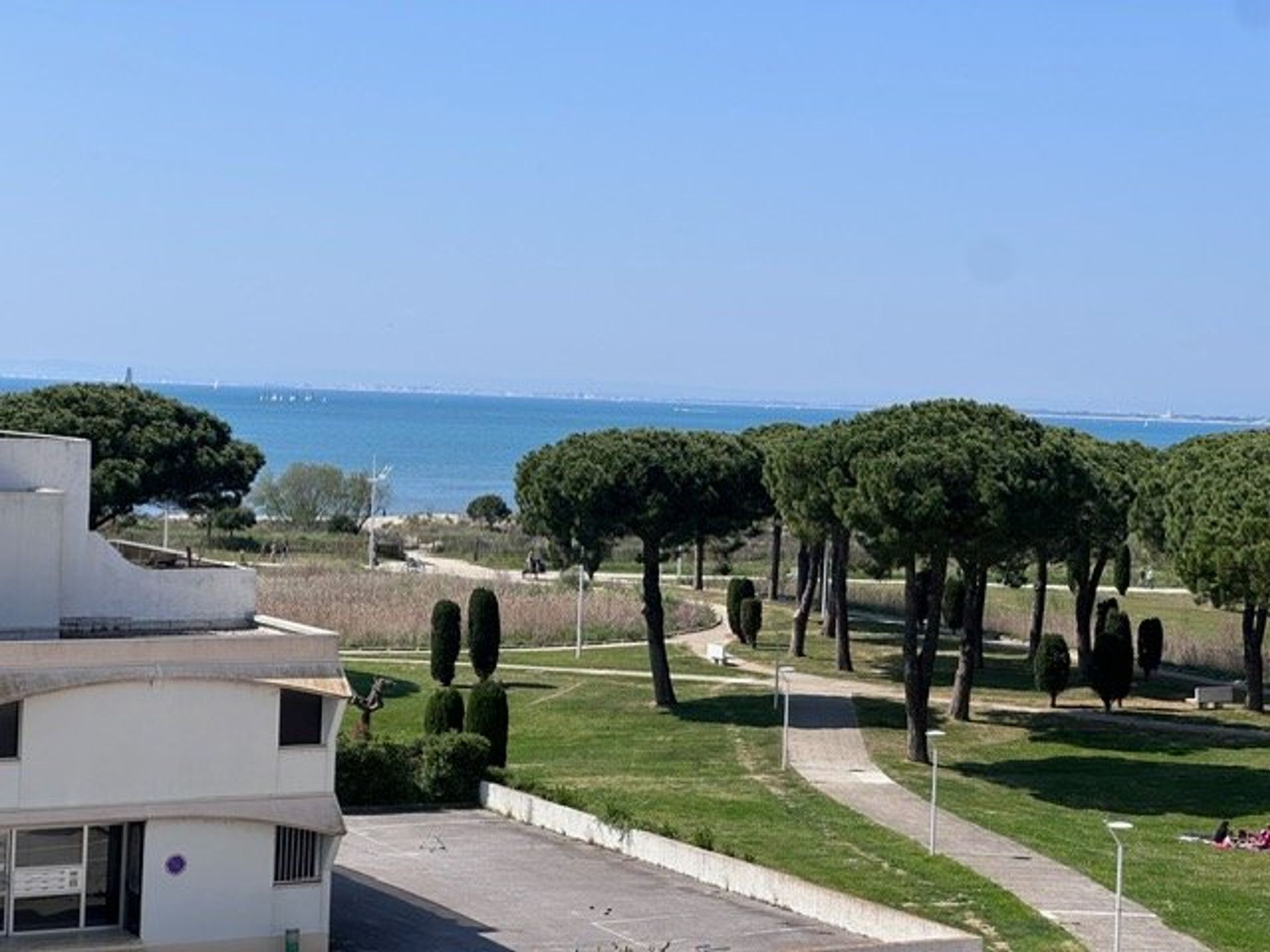 Un bâtiment doté d’un toit plat et d’une zone pavée devant l’entrée. On aperçoit des arbres et un ciel dégagé en arrière-plan.