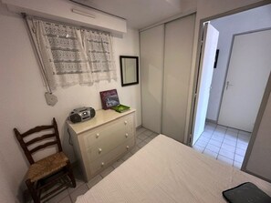 Un dormitorio con cama, cómoda, silla, radio, cuadro enmarco y puerta que da a otra habitación.