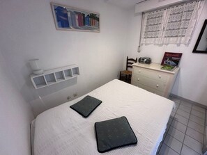 Une chambre à coucher avec un lit, une table de chevet avec une radio, une chaise et une fenêtre ornée de rideaux en dentelle.