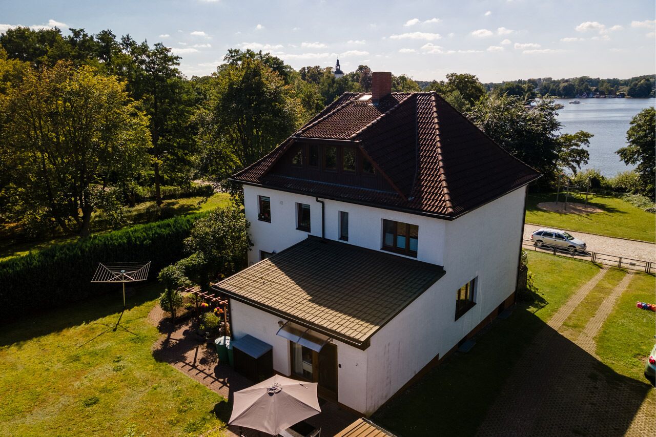 A white house with a sloped roof and a small porch, surrounded by lush greenery and a lake.