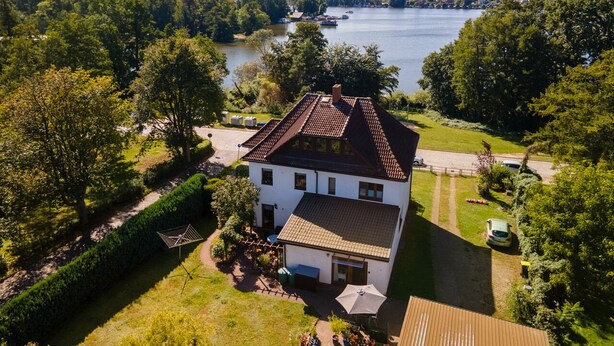 A two-story house with a tiled roof, surrounded by a well-maintained garden and a view of a lake with boats. - Hochwertiges 1-zi-app für 2+1 Pers. am See, Wlan, Terrasse, Elektr. Kamin (Mirow)