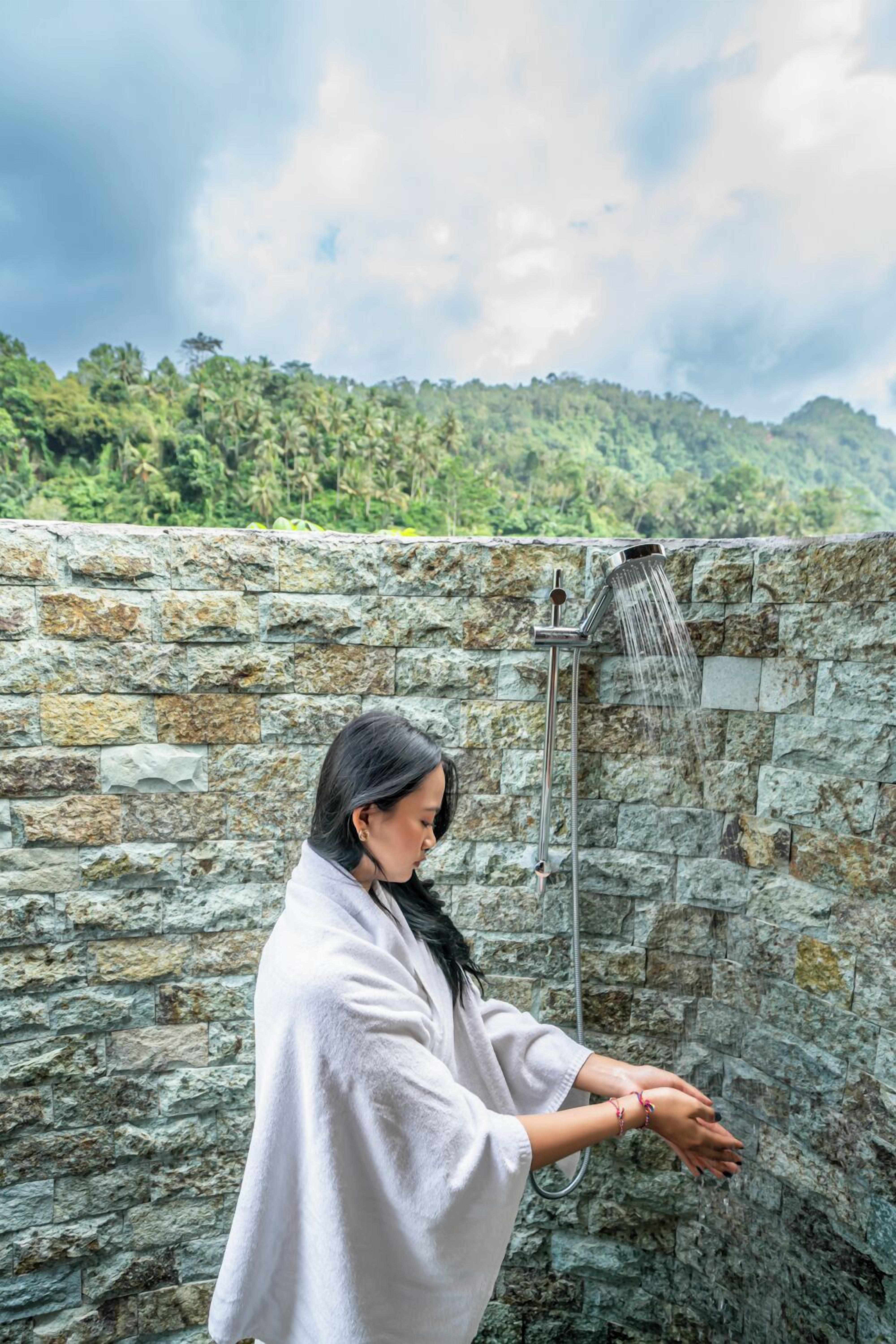 A person in a white robe standing under a shower in a stone-walled room with a view of a green hillside.