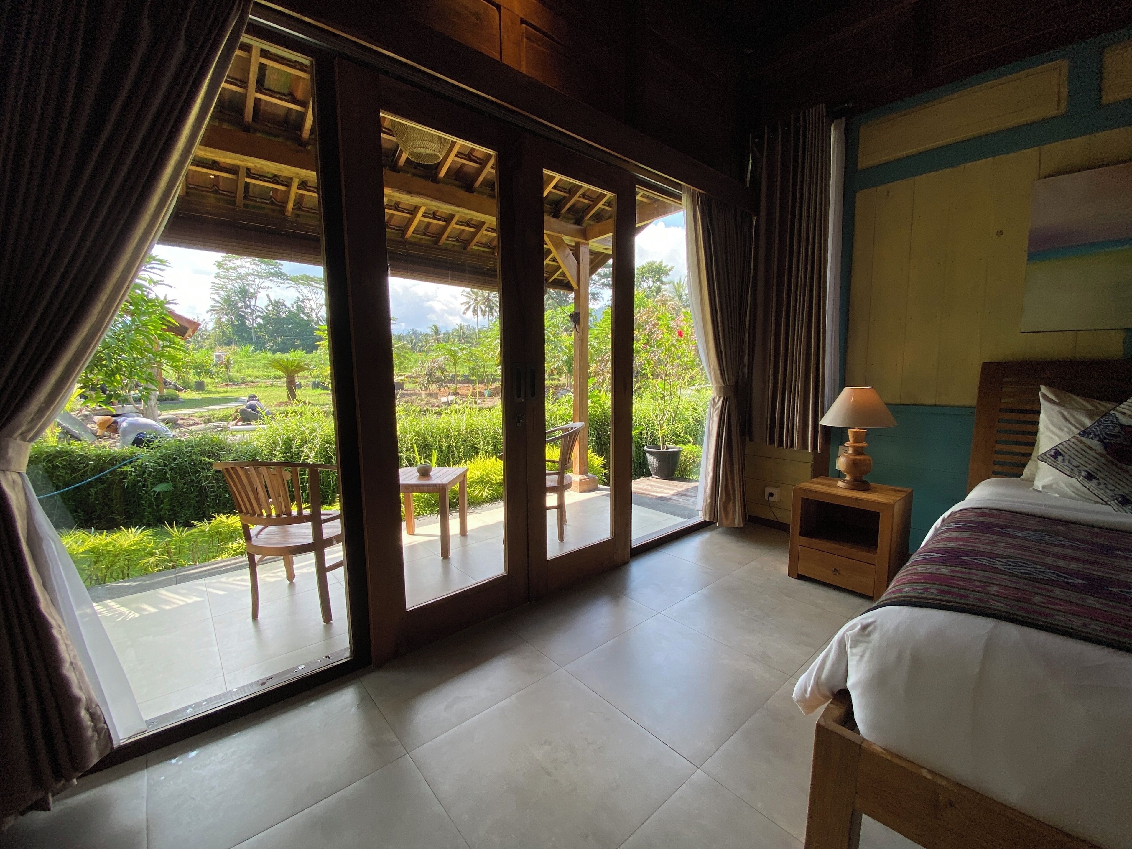 A room with a bed, a small table with a lamp, and a view of a garden through a sliding door.