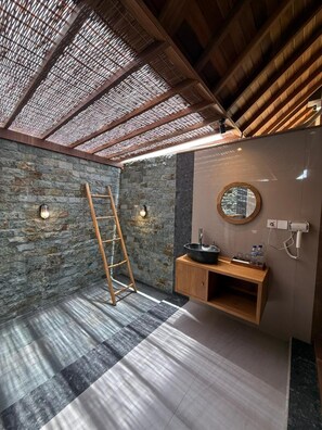A bathroom with a stone wall, a wooden ladder, a round mirror, a wooden vanity, and a black sink.