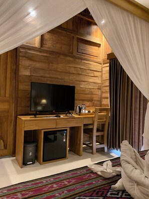 A cozy room with a wooden desk, a flat-screen TV, a small refrigerator, and a chair.