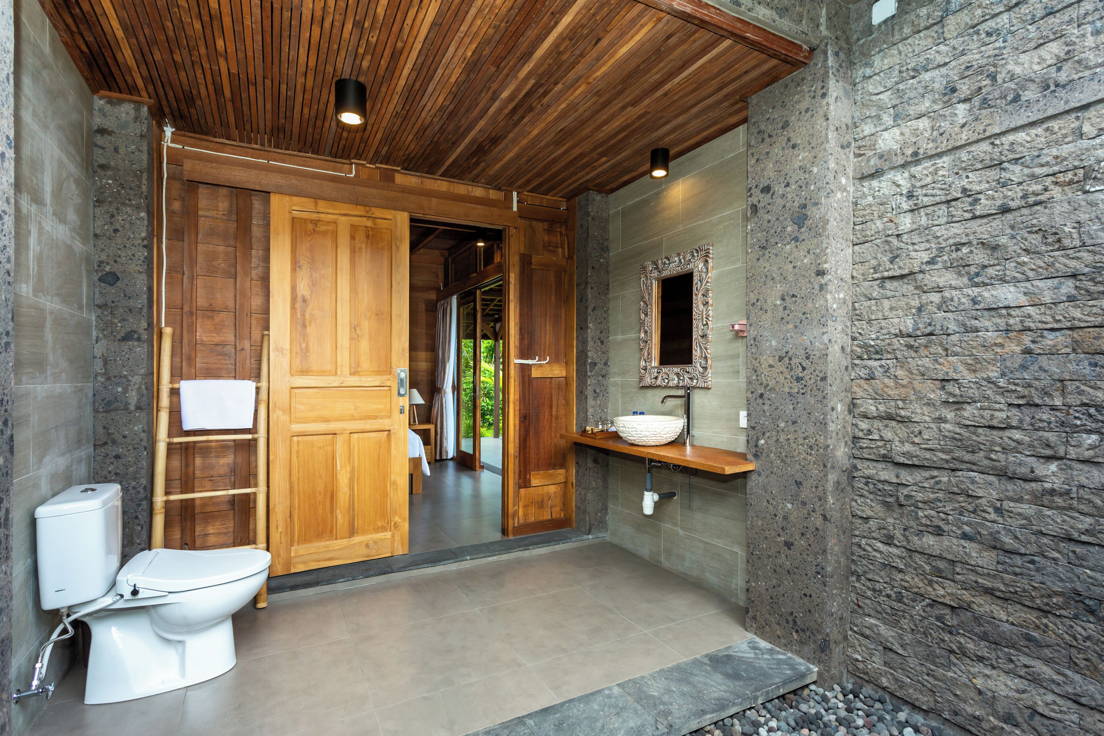 A modern bathroom with a wooden door, a white toilet, a stone wall, and a mirror above the sink.