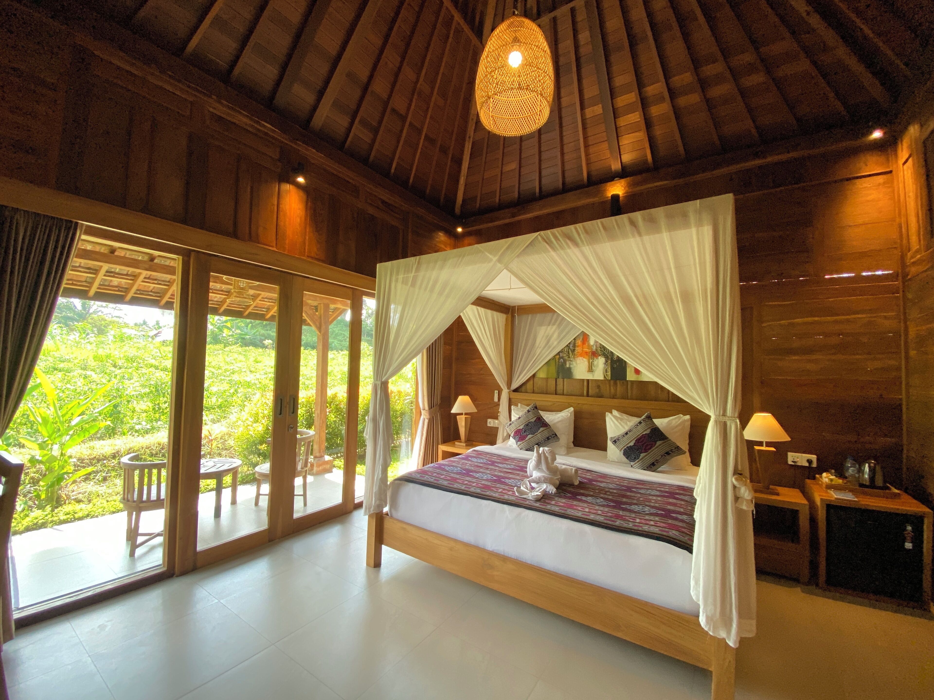 A spacious bedroom with a large bed, a canopy, and a view of a lush garden through sliding glass doors.