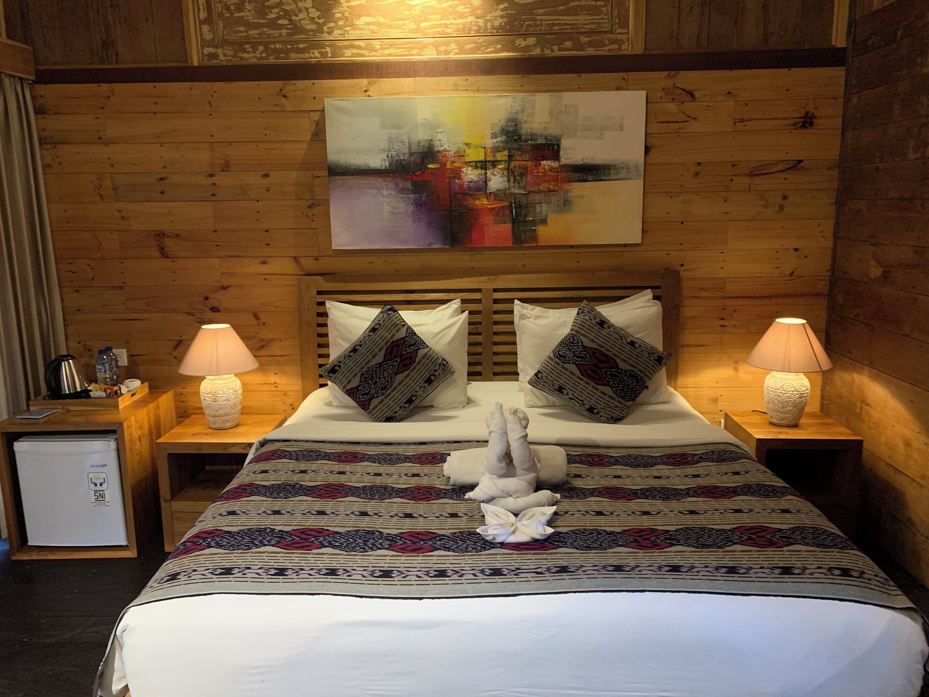 A wooden-paneled room with a bed, two bedside lamps, a painting above the bed, and a small refrigerator on the left.