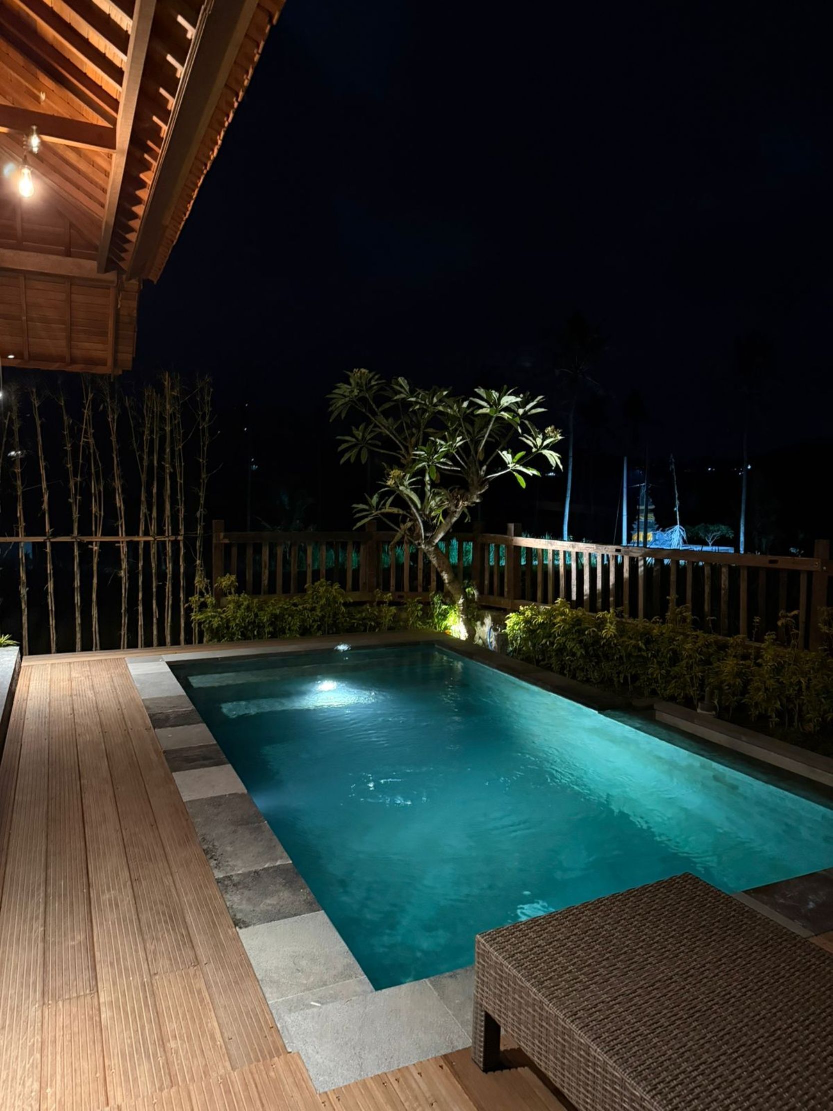 Una piscina con cubierta de madera, rodeada de bambú y plantas tropicales, bajo un cielo nocturno.