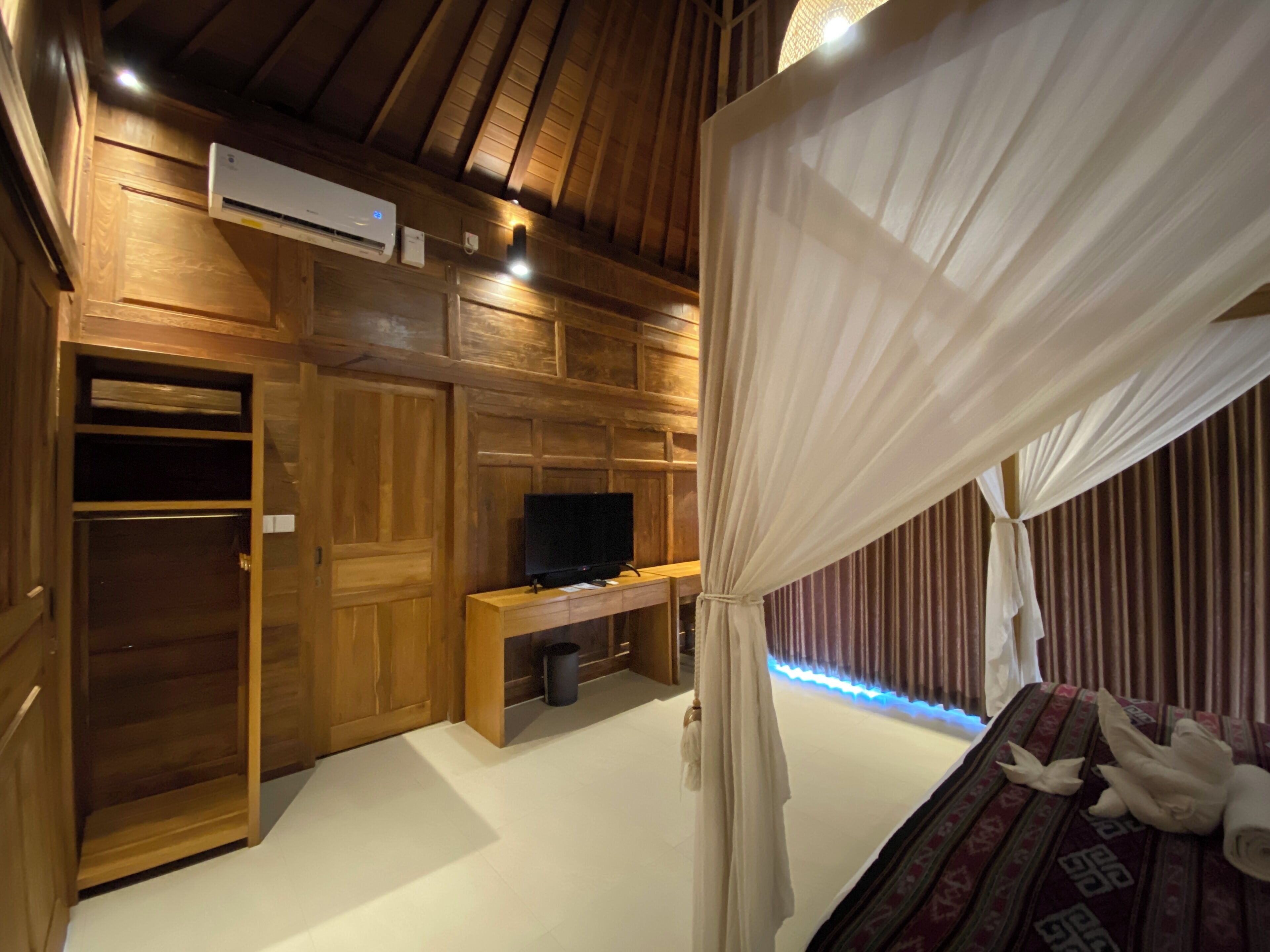 Ein Zimmer mit Holzbalkendecke, einem Fernseher auf einem Holzboden und einem Bett mit Baldachin, sowie einer Wand-Klimaanlage.