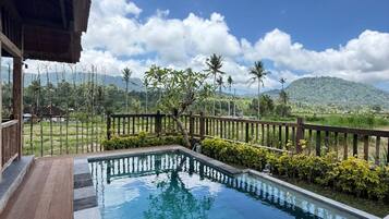A swimming pool with a wooden deck, surrounded by a wooden fence and lush greenery, with a mountain in the background.