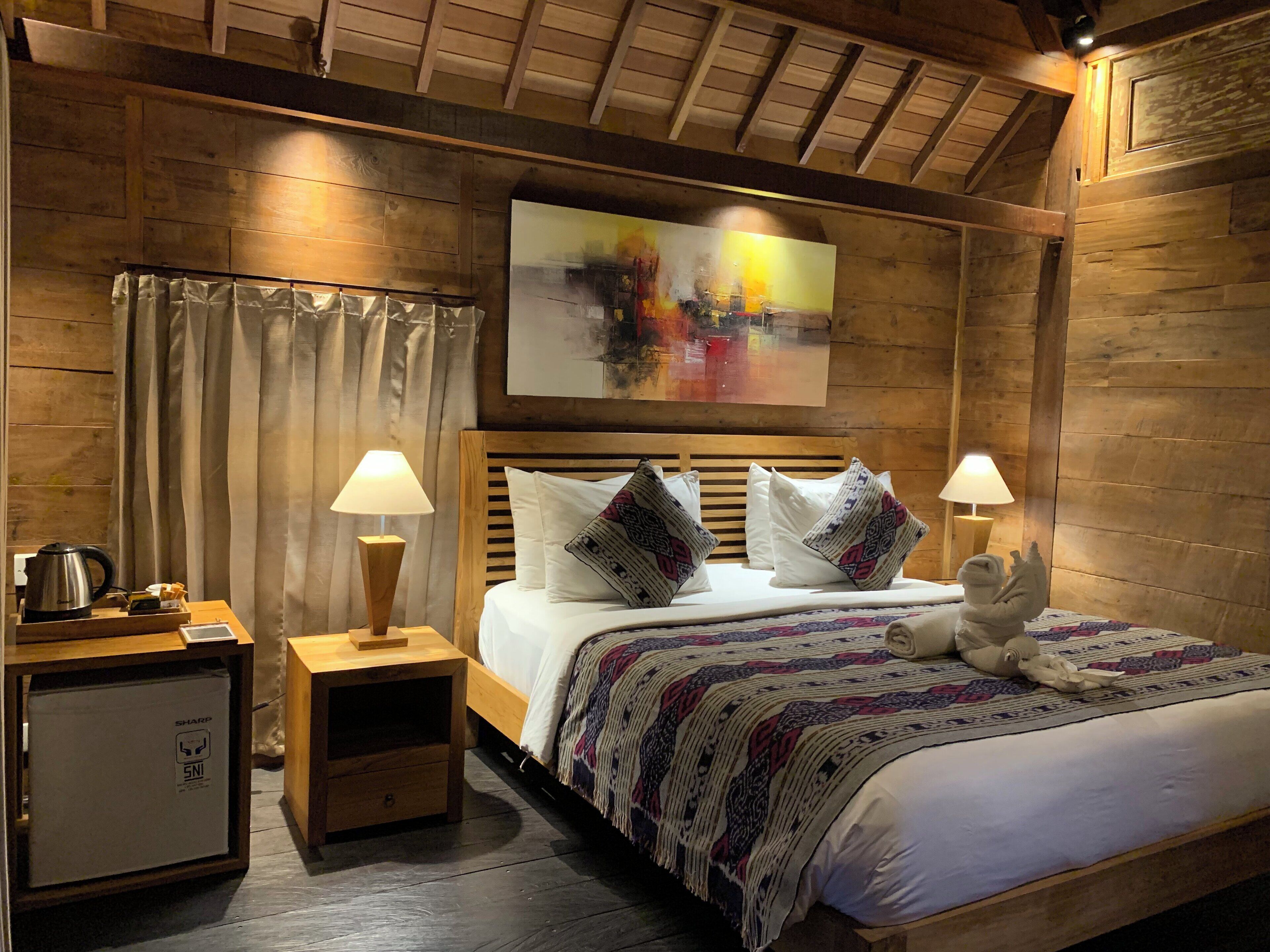 A wooden room with a bed, two bedside lamps, a small table with a kettle, and a painting on the wall.