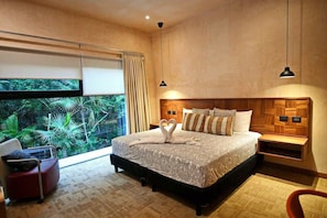 Une chambre avec un grand lit, une tête de lit en bois, une table de chevet, une chaise et une vue sur la verdure à l’extérieur.
