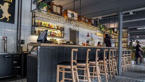 Zona de bar con suelo de baldosas, sillas de madera, barra con caja registradora y estantes llenos de botellas.