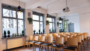 Un salón de conferencias con filas de sillas de madera, ventanas amplias con plantas en macetas y lámparas colgantes.