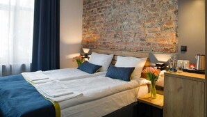 Habitación de hotel con pared de ladrillo decorativa, cama con ropa blanca, mesita de noche con un florero y escritorio de madera con tetera.