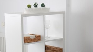 A white shelving unit with wicker baskets and glassware, a gray sofa with a blue pillow, and a fern plant. - Paseo Maritimo (Málaga)