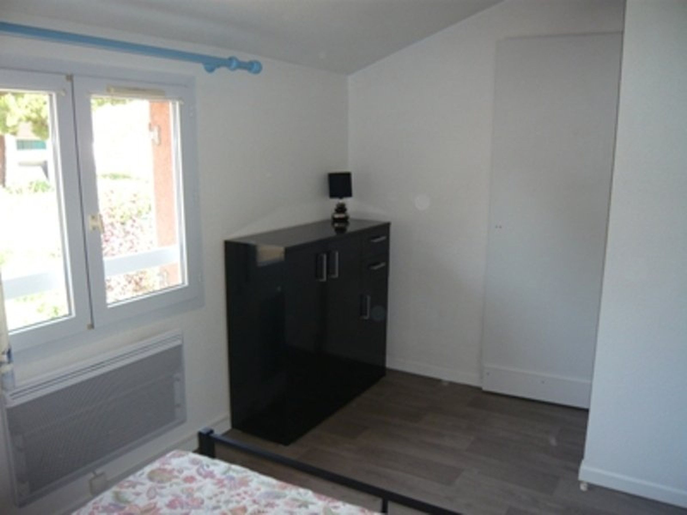 Ein Zimmer mit einem Bett, einem Fenster, einer Heizung, einem schwarzen Schrank und einer Tür.