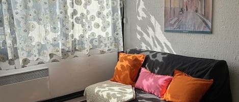 Una habitación con sofá, ventana con cortinas florales, radiador y un cuadro en la pared.