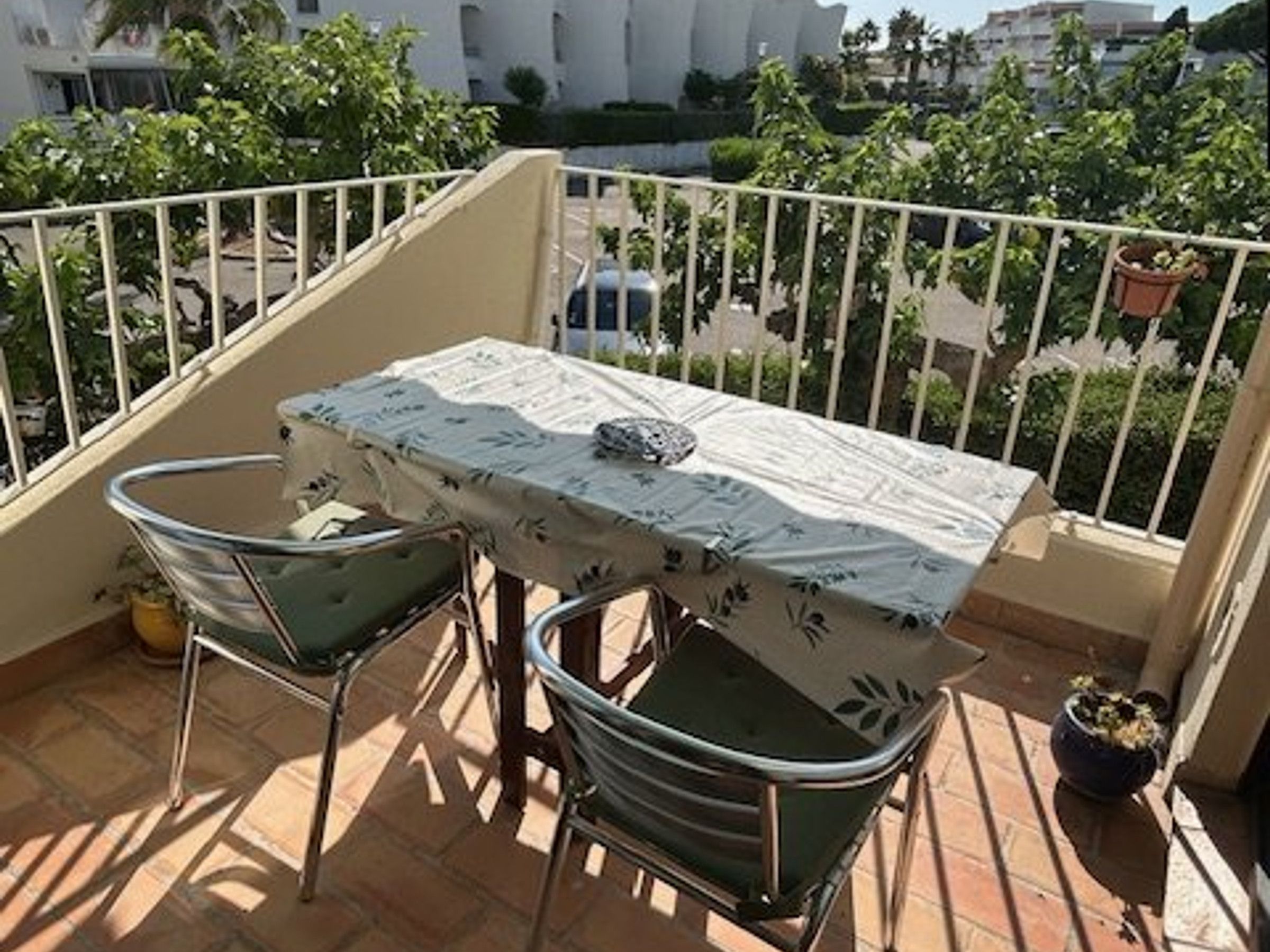 Varanda com mesa e duas cadeiras, planta em vaso e vista para edifícios e vegetação.
