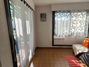 A room with a sliding glass door, a window with floral curtains, a sofa with an orange cushion, and a red and white striped rug. - Cozy 1BR Apartment with Terrace, Parking, Next to Beach and Shops (Port Camargue)