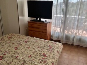 A bedroom with a bed, a television on a wooden dresser, and a window with sheer curtains. - Cozy 1BR Apartment with Terrace, Parking, Next to Beach and Shops (Port Camargue)