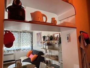 Een knus woongedeelte met een kitchenette, een bank met kussens, een raam met gordijnen en een plank met diverse spullen.