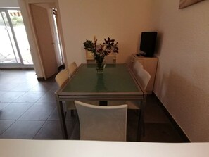 A dining area with a glass-top table, white chairs, a vase with flowers, and a television on a stand. - 2-room apartment with private parking, 600m from shops (Port Camargue)
