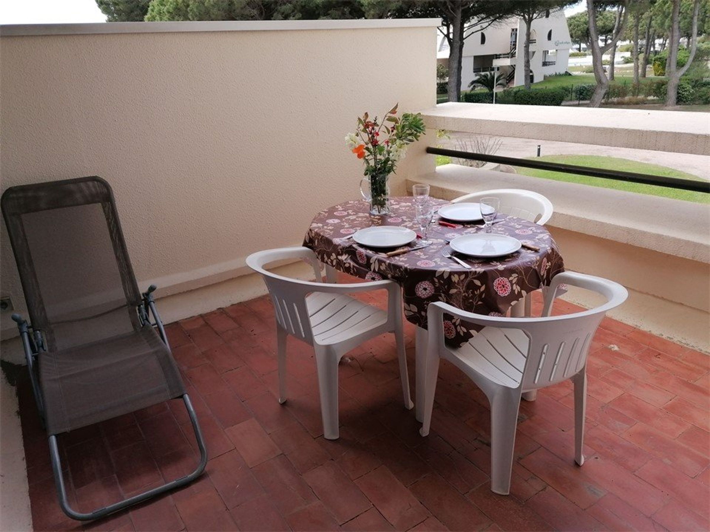 A terrace with a table set for two, a folding chair, and a view of a building and trees.