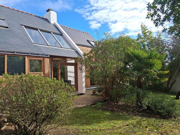 Une maison avec un toit en pente, des fenêtres de toit et un jardin où poussent différentes plantes, ainsi qu’un palmier.
