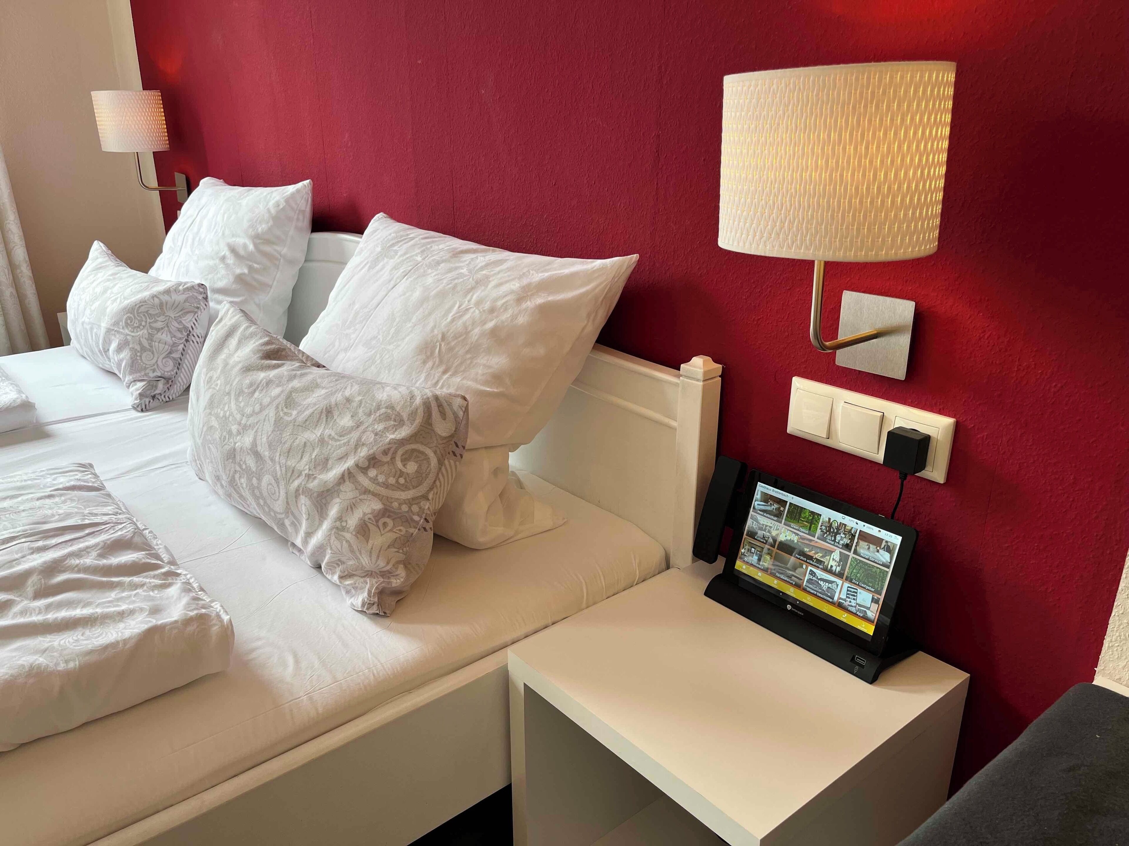 A hotel room with a bed, pillows, a bedside table with a lamp, a wall-mounted TV, and a red accent wall.