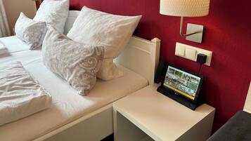 A hotel room with a bed, pillows, a bedside table with a lamp, a wall-mounted TV, and a red accent wall.