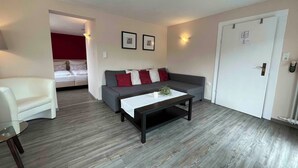 Una sala moderna con un sofá gris, un sillón blanco y una mesa de centro. Hay una puerta con cerradura y una lámpara de pared.