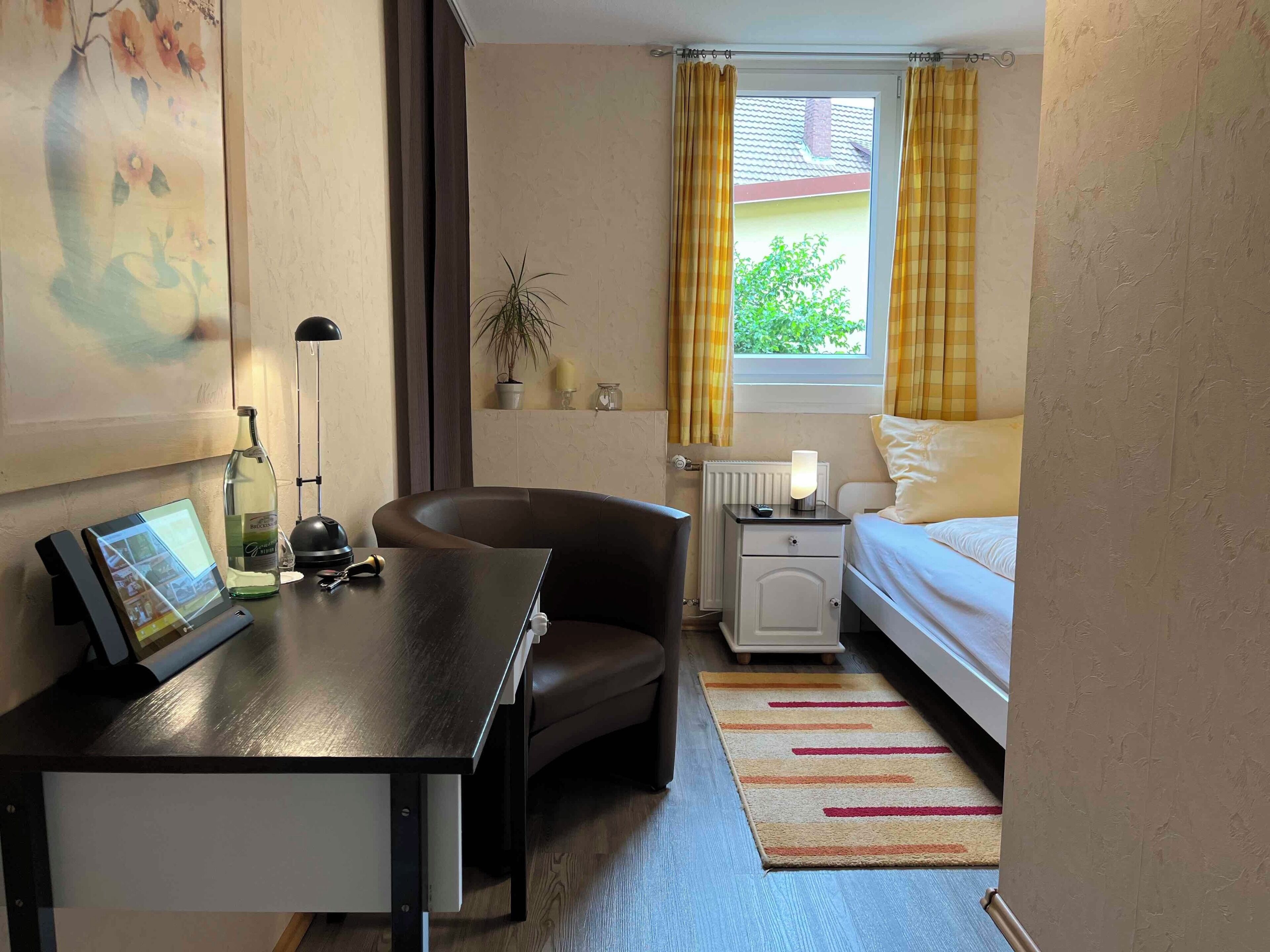 Habitación de hotel con cama, escritorio con computadora, silla, mesita de noche con lámpara y ventana con cortinas.