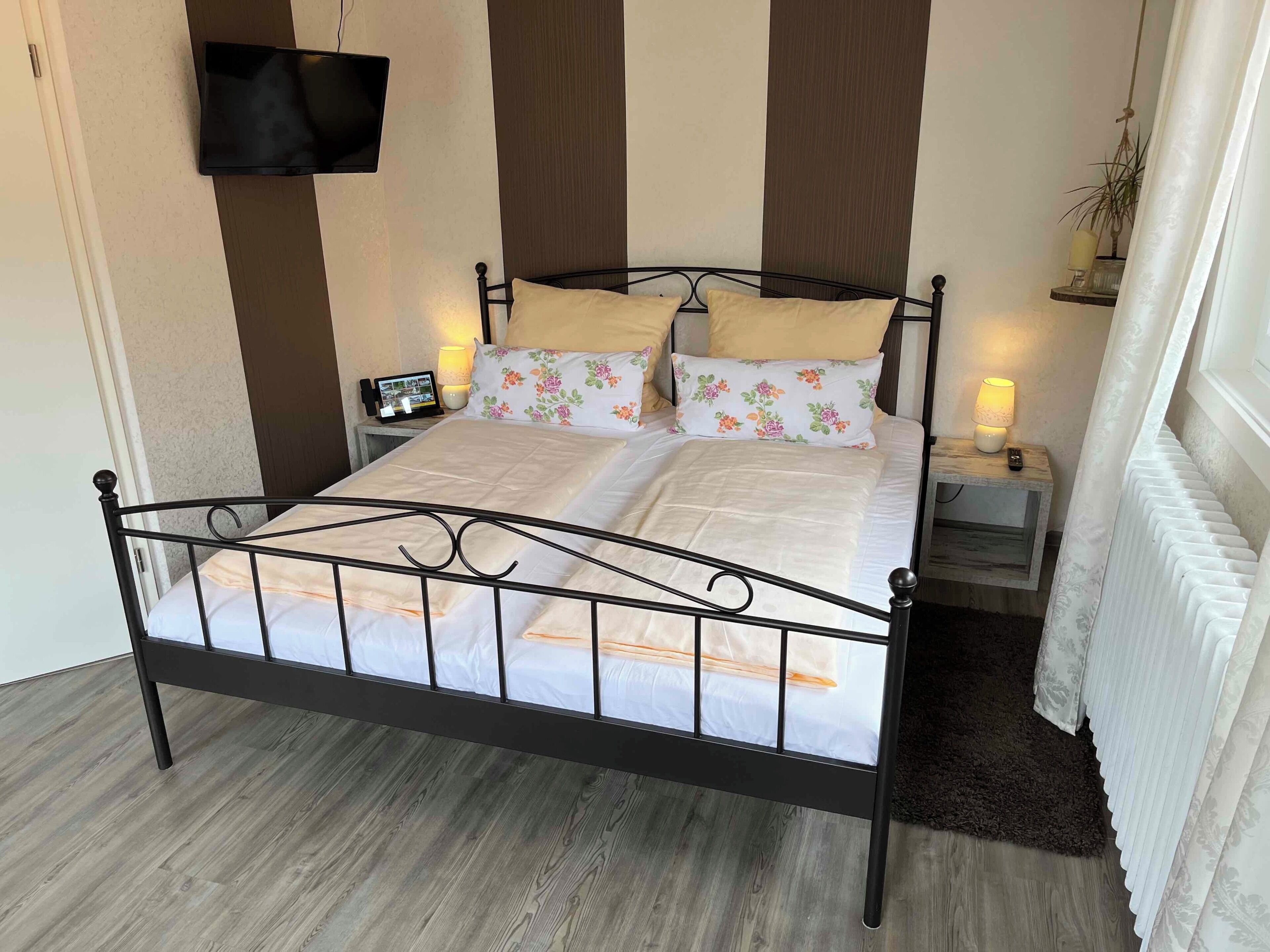 A neatly made bed with a metal frame, two bedside tables with lamps, a wall-mounted TV, and a radiator.