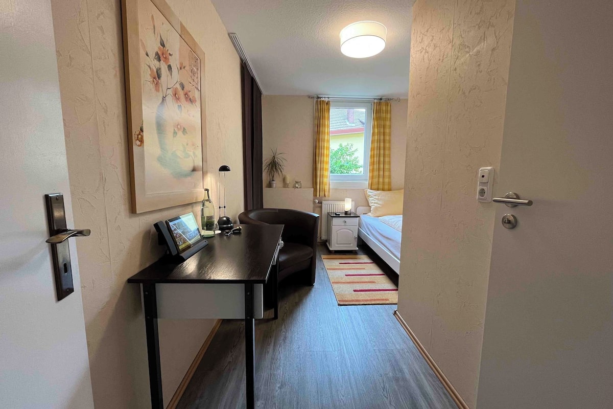 Hotellihuone, jossa on työpöytä, sänky, yöpöytä, lamppu, valokuvakehys ja ikkuna, jossa on verhot.