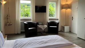 Habitación de hotel con cama, dos sillones, televisor, una mesita con una computadora portátil y ventanas amplias con cortinas.