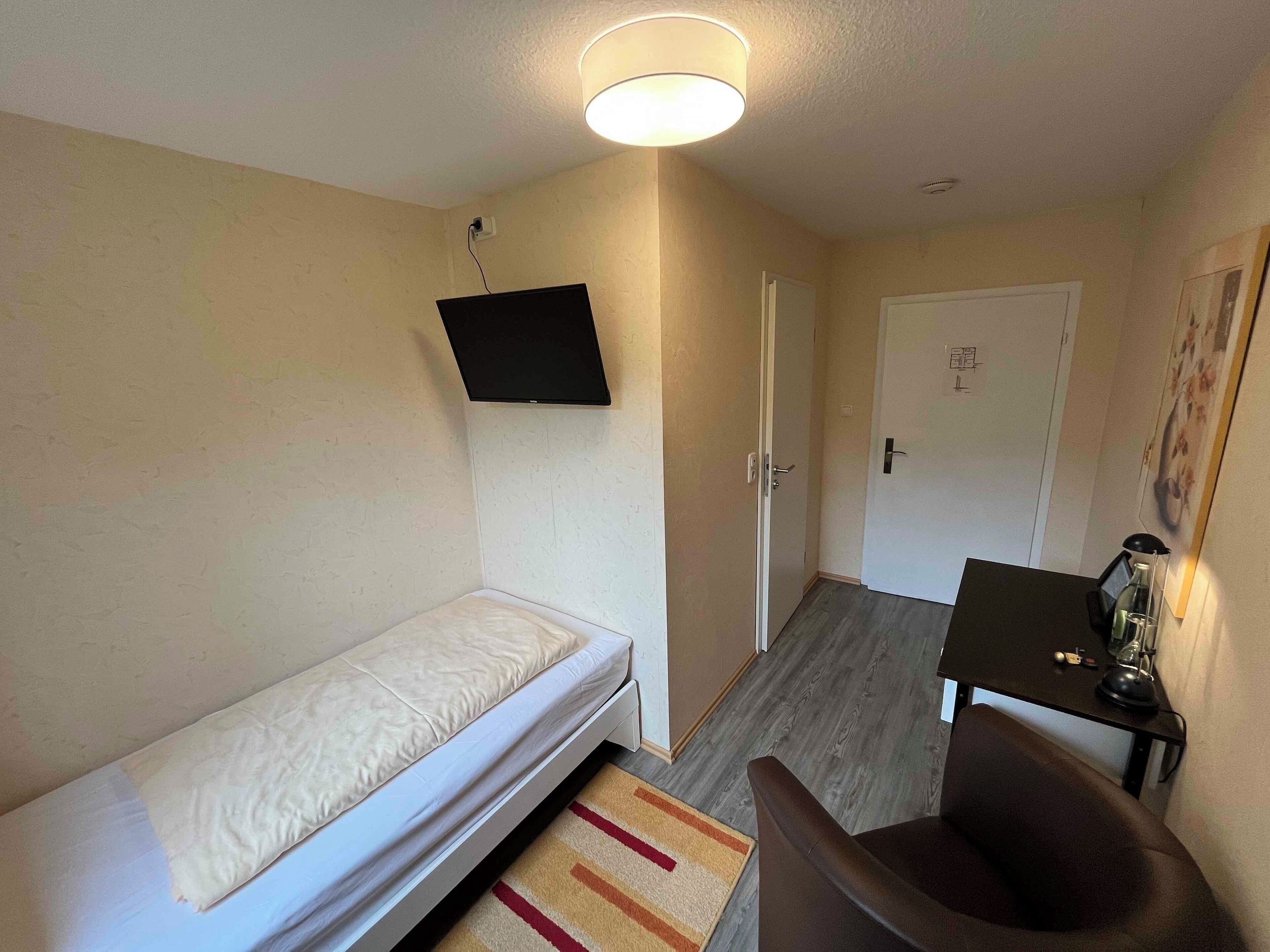 Una habitación pequeña y funcional con una cama individual, un escritorio con silla, un televisor montado en la pared y una puerta que da a otra habitación.