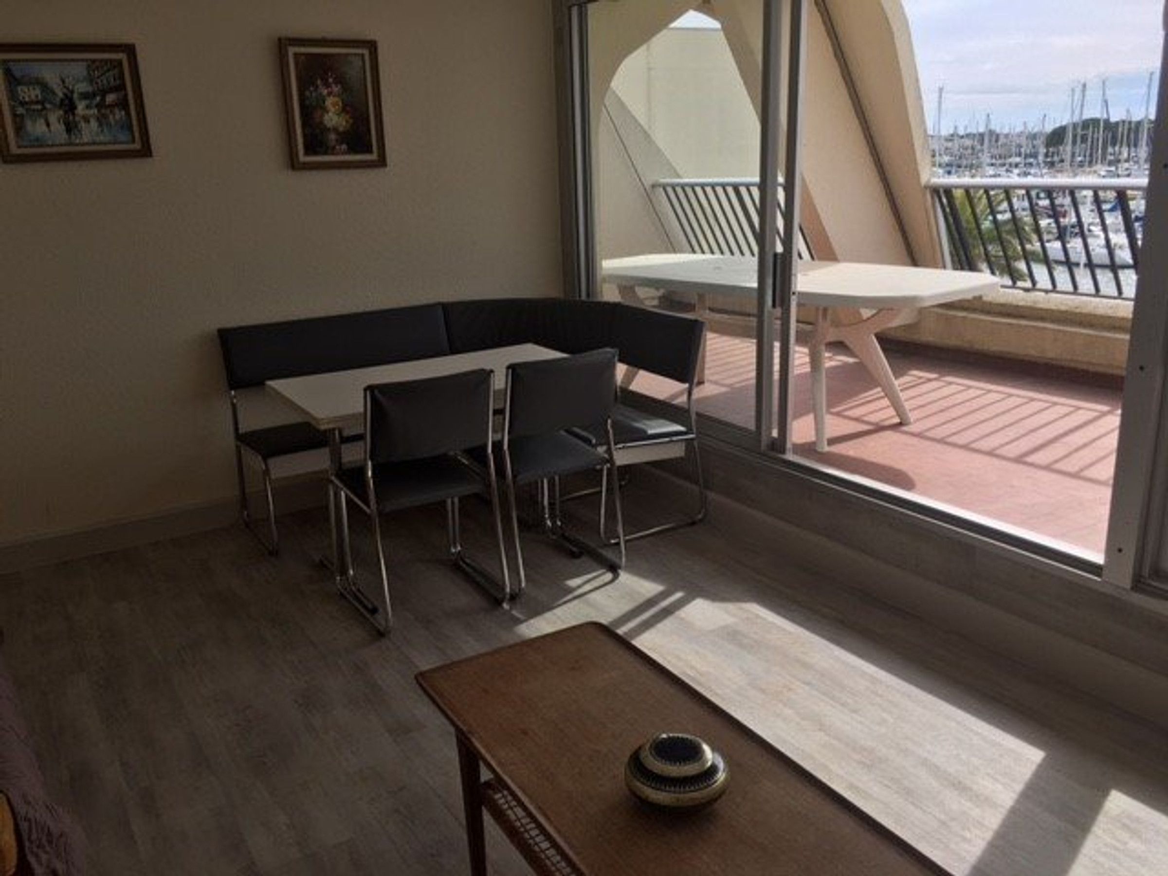 Une pièce comprenant une table à manger et des chaises, un canapé, une petite table avec un saladier, et une vue sur une marina grâce à de grandes fenêtres.