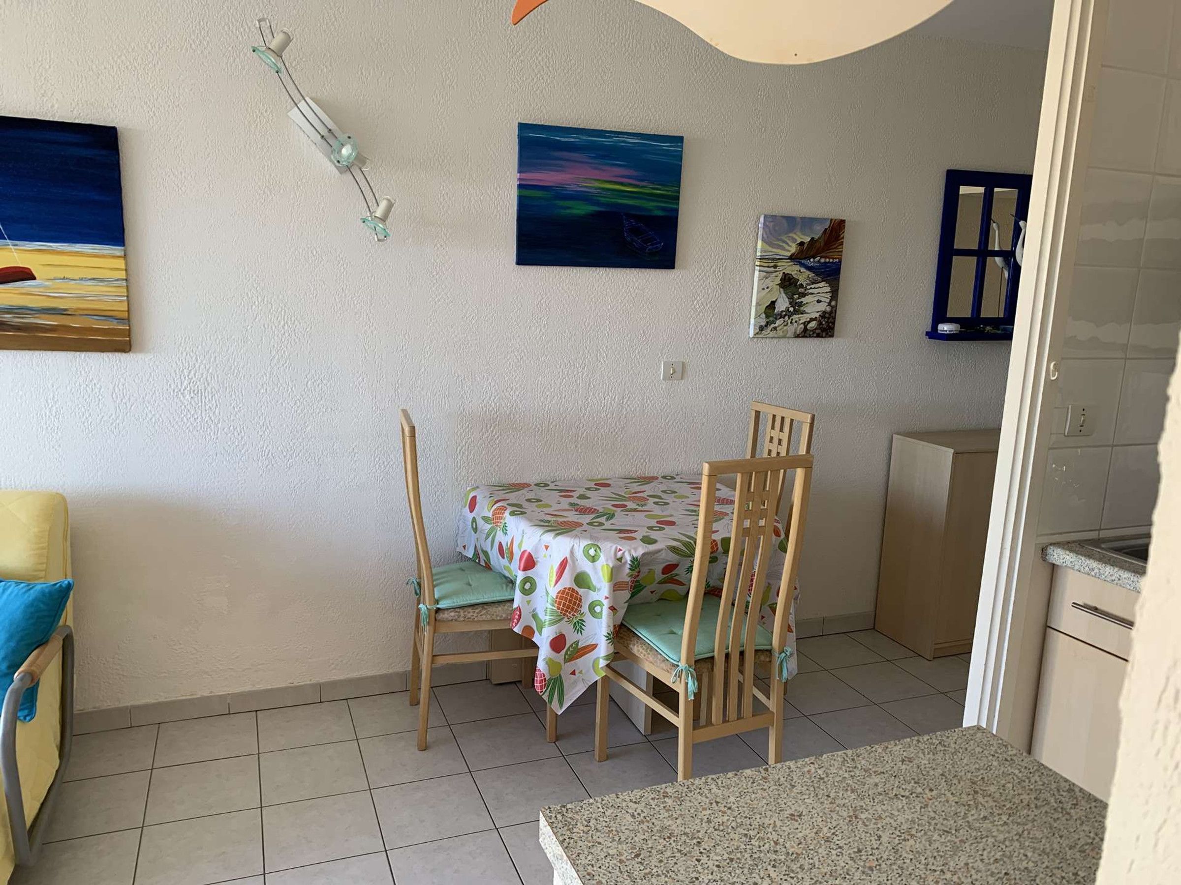 Un petit coin repas avec une table et des chaises, un espace cuisine et deux tableaux accrochés au mur.