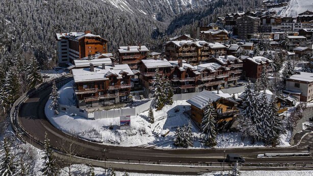 Un centro turístico de montaña cubierto de nieve, con edificios de madera, un telesilla y una carretera serpenteando por la zona.