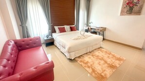 Une chambre d’hôtel avec un lit, une chaise rouge, une tête de lit en bois, une table de chevet, un bureau et un tableau accroché au mur.