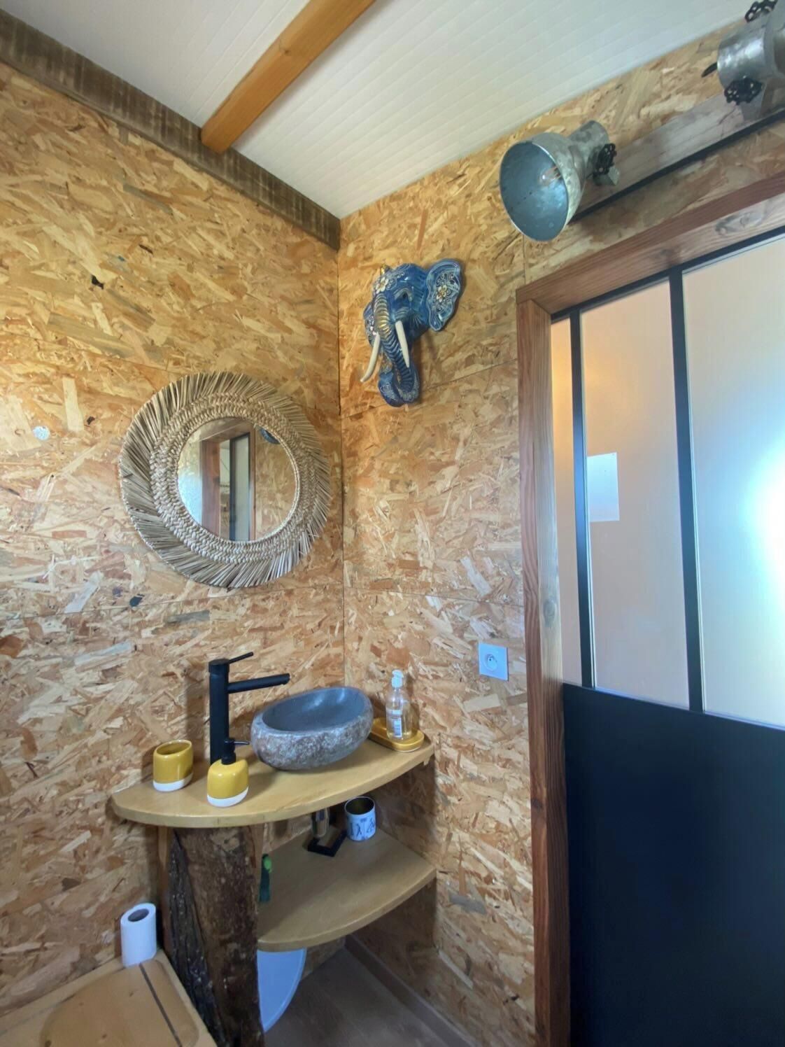 Ett badrum med en väggpanel av trä, en rund spegel, ett handfat med en skål av sten och ett dekorativt elefantansikte.
